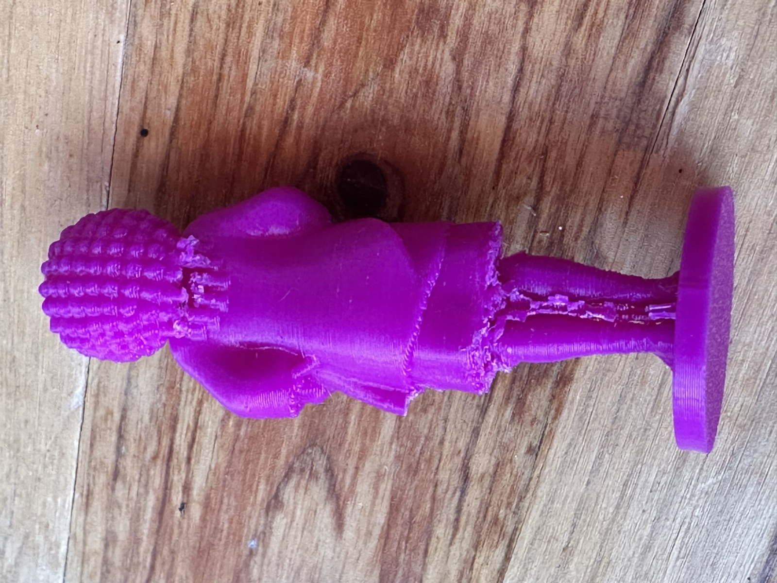 A purple 3D-printed figurine seen from behind on a wooden table: textured hair, a jacket, a rough hem and marks down the calves where supports were removed