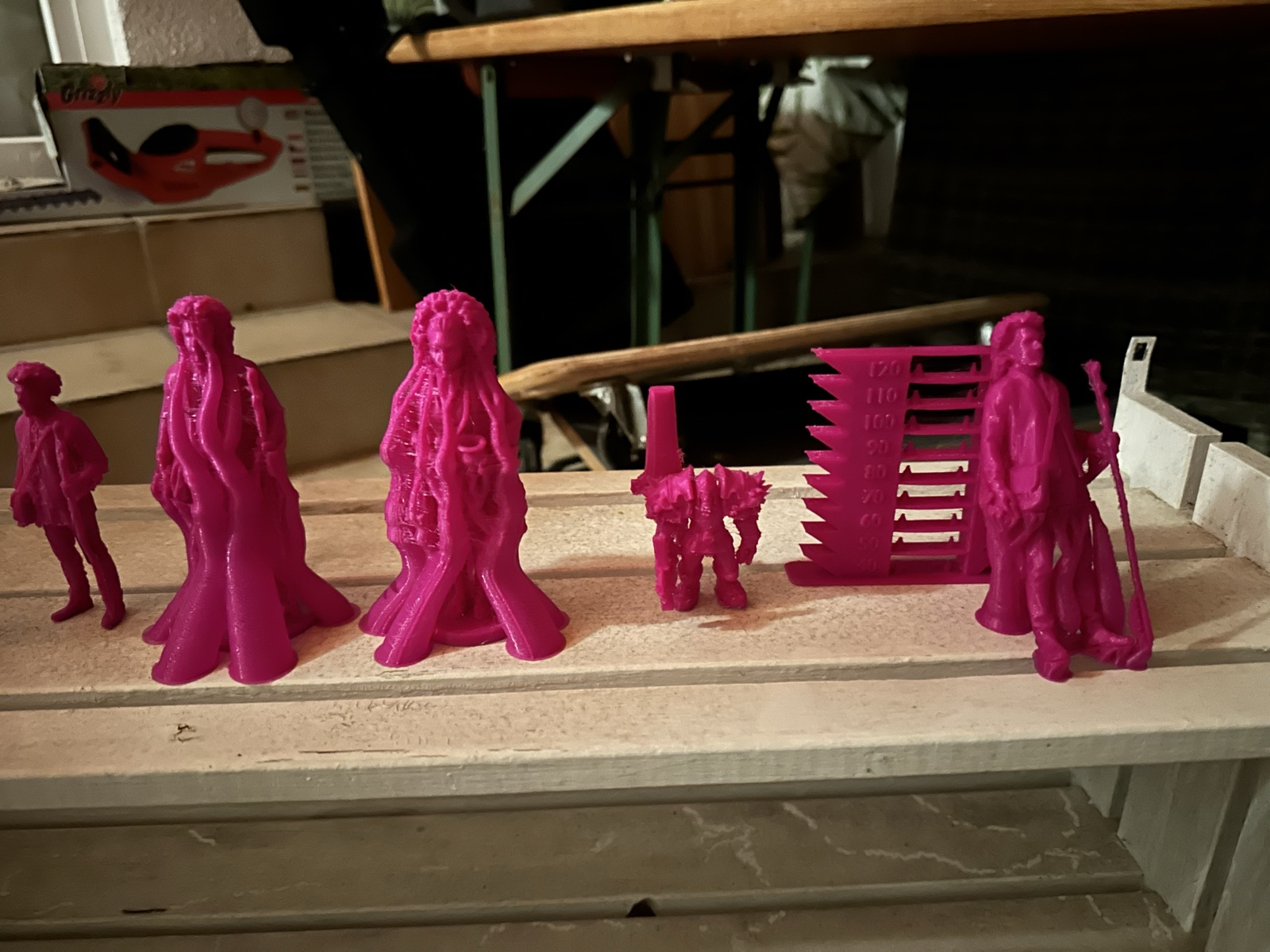 Six magenta 3D prints in a row on a wooden bench: a small walker, two Amara figurines still wrapped in support strands, a small orc, a stepped temperature tower marked 40 to 120, and Daniel with a staff