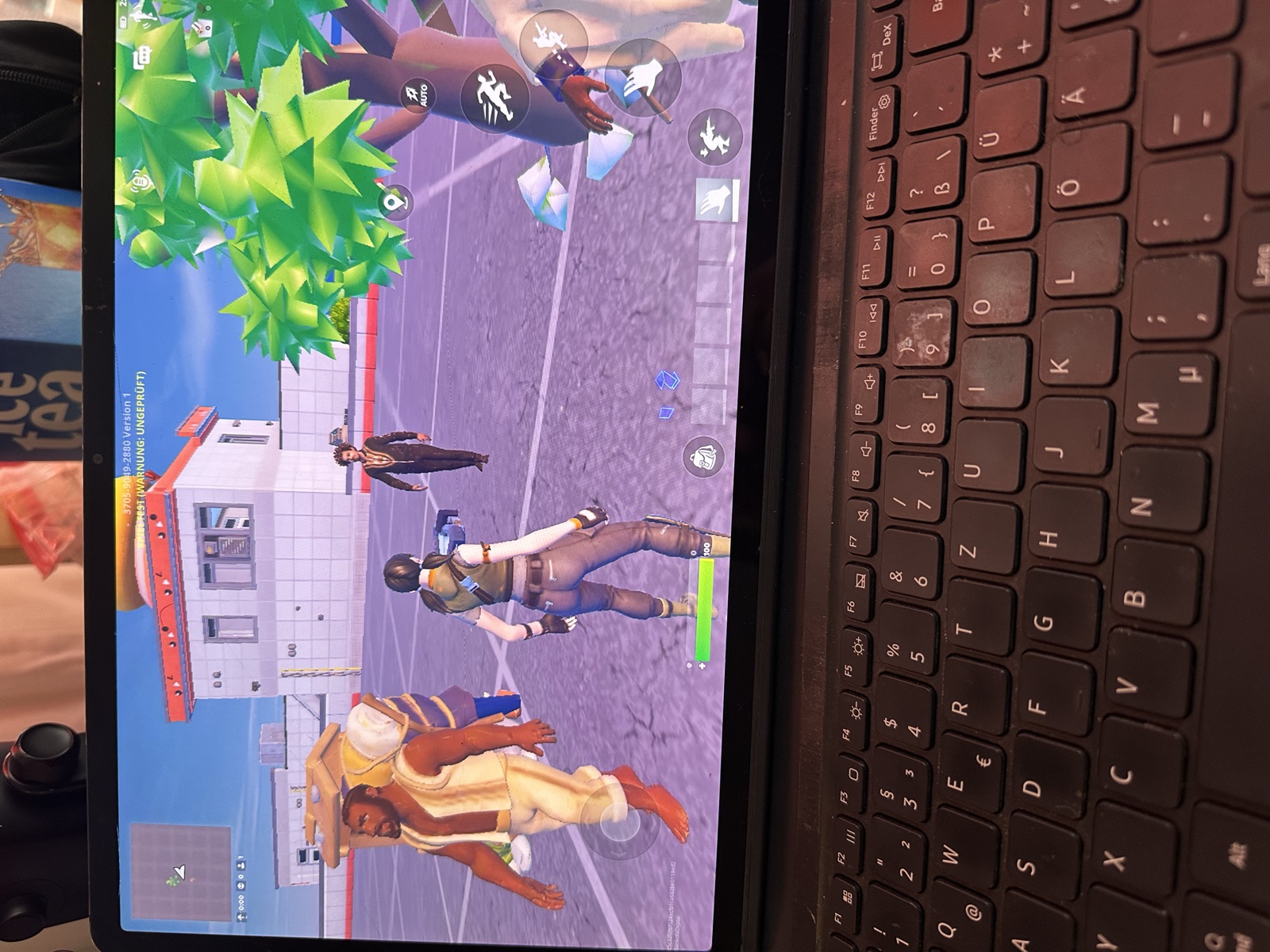 A tablet on a keyboard showing a Fortnite test island: the carrier with a large pack on his back and the walker in a dark coat stand on a grey plaza beside a Fortnite avatar, a white building and a stylised tree behind them